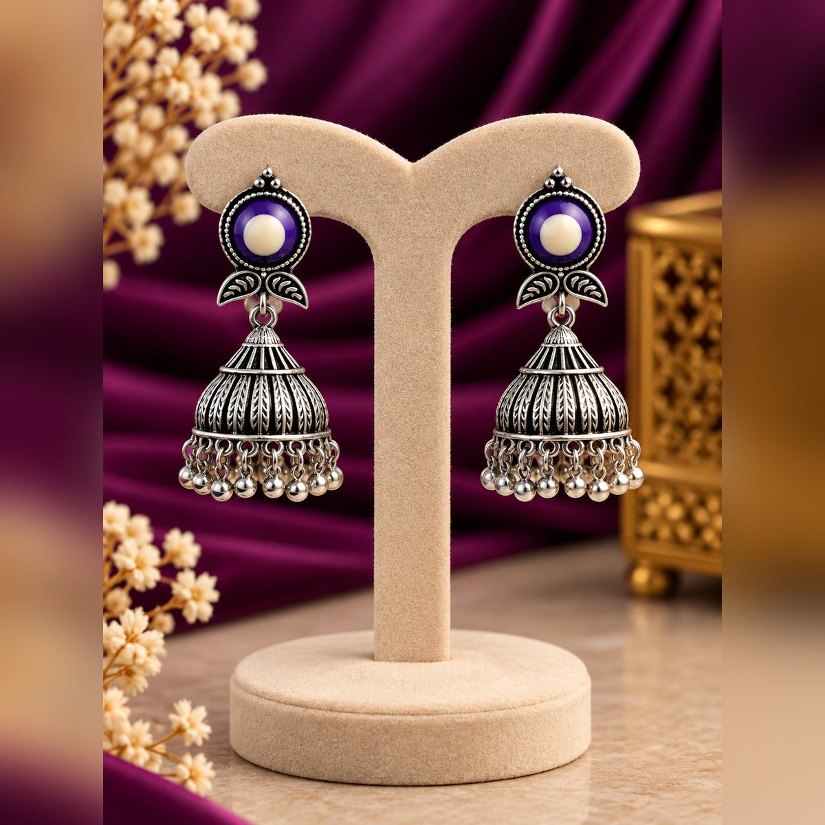 Oxidized silver jhumka earrings with purple and white evil-eye studs and ribbed dome bells with ball fringe, on a display stand