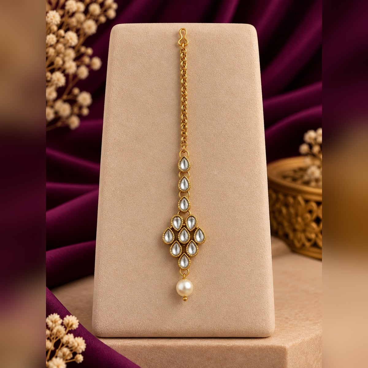 Gold-tone maang tikka with teardrop Kundan stones and a pearl drop, on a display stand